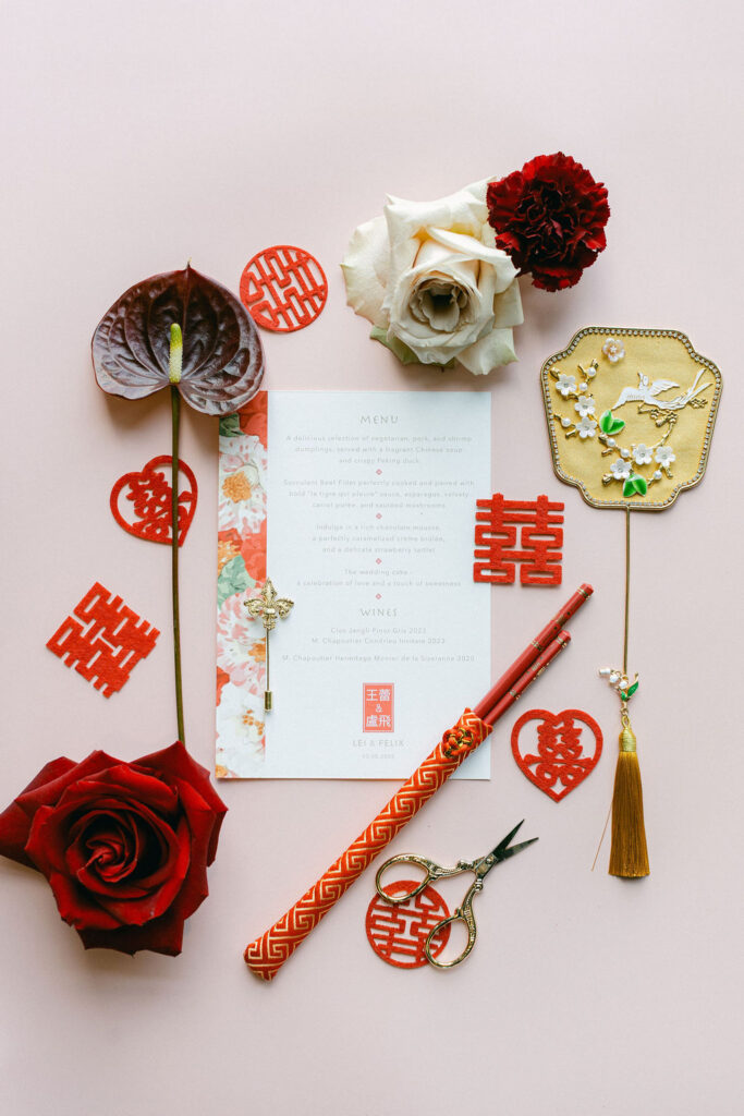 Chinese wedding flat lay with double happiness symbols, red scissors, a calligraphy pen, anthurium, roses, and a handwritten ceremony card