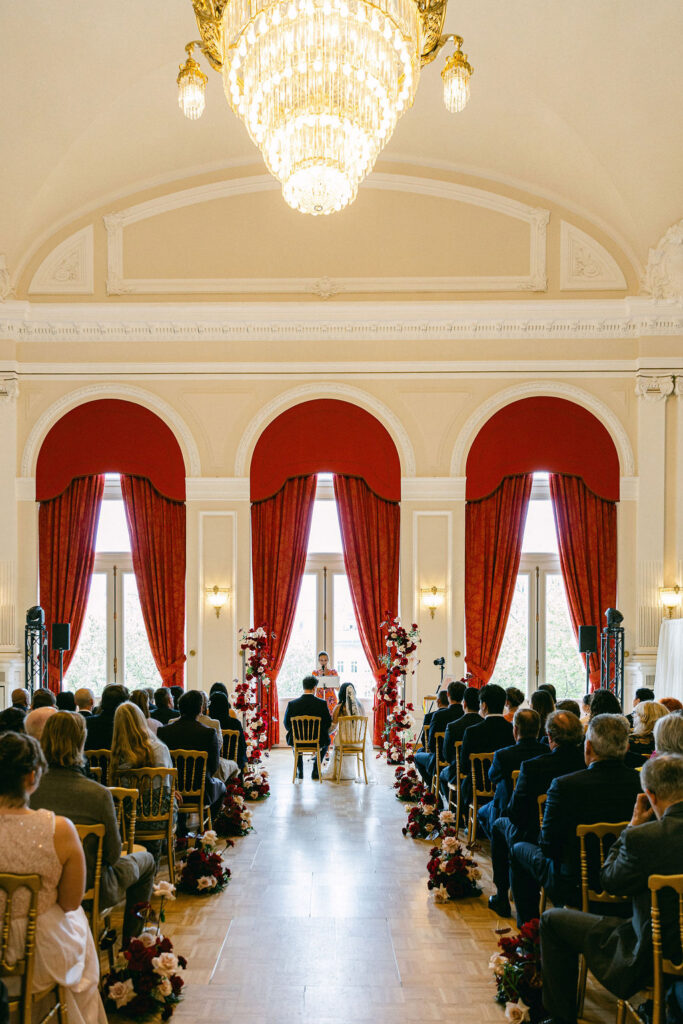 Wedding ceremony at Cercle Cité in Luxembourg City, with guests seated in gold chairs along a floral aisle leading to a couple at the altar, framed by dramatic red draped arches and a grand crystal chandelier