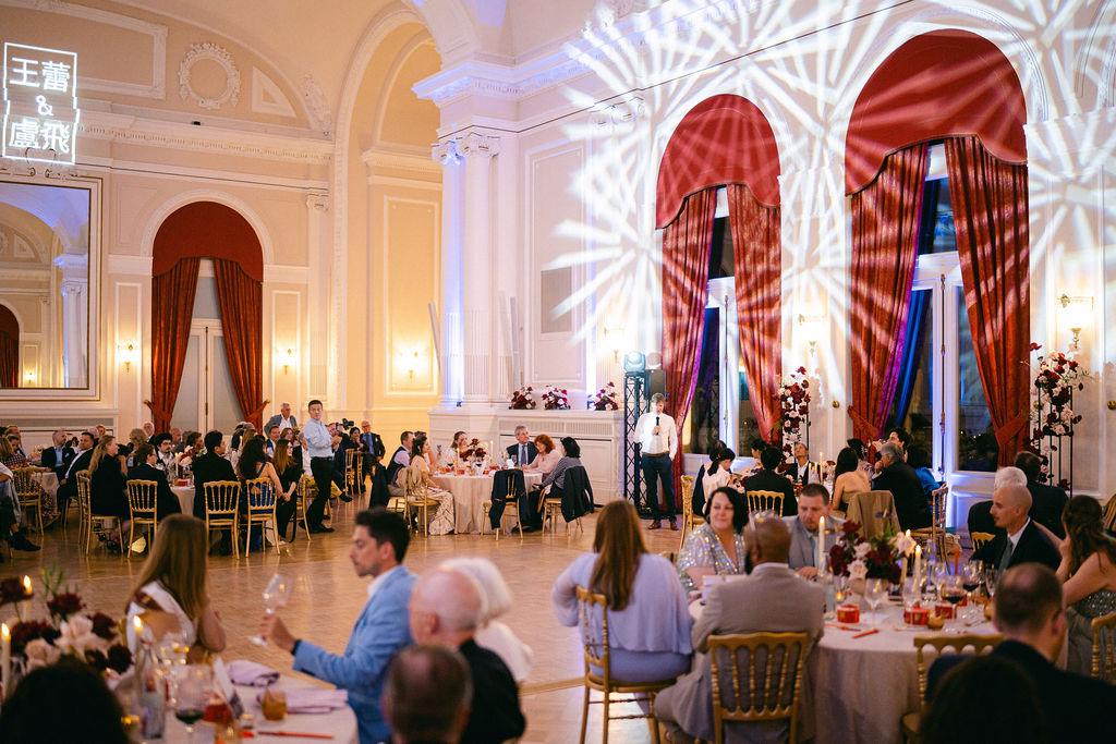 Wedding reception in full swing at Cercle Cité Luxembourg with guests dining under dramatic projected lighting, Chinese character monogram signage, and tall floral installations