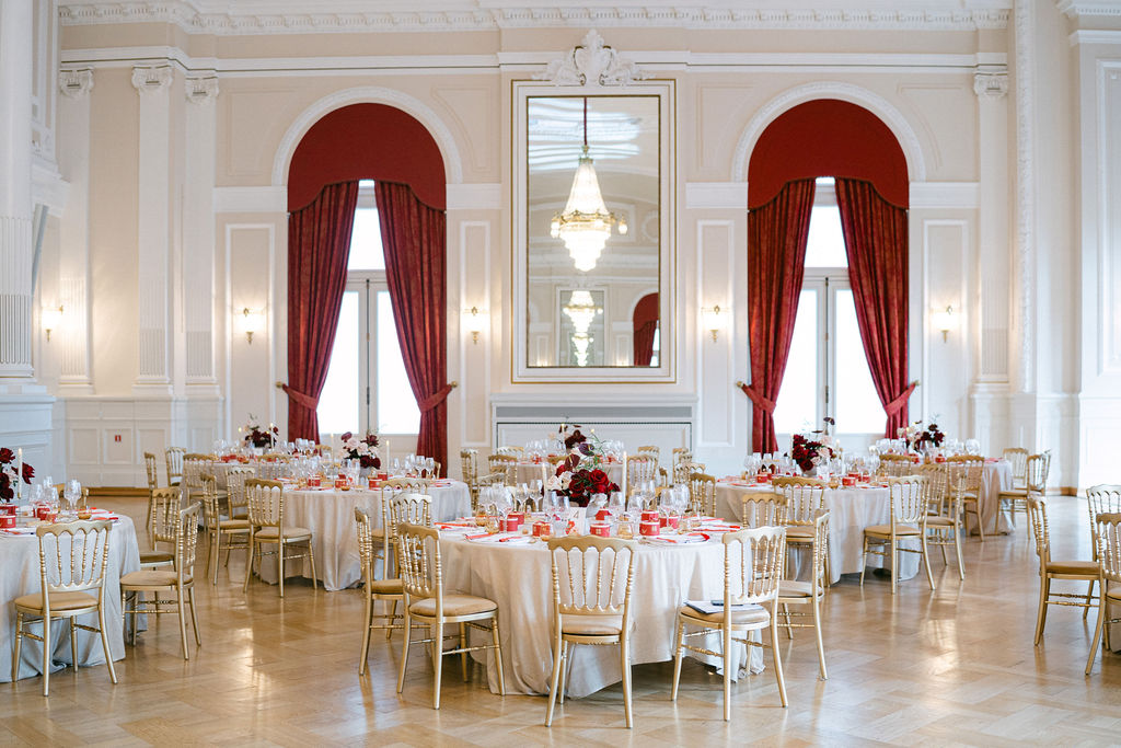 Wedding reception room at Cercle Cité Luxembourg fully set with round tables, gold Chiavari chairs, deep red floral centerpieces, and dramatic red draped arches