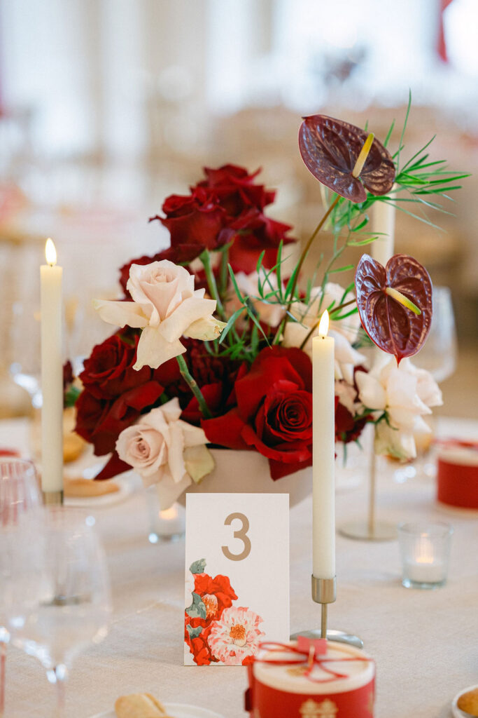 Luxury wedding reception table detail at Cercle Cité featuring deep red roses, anthurium blooms, taper candles, and a hand-lettered table number
