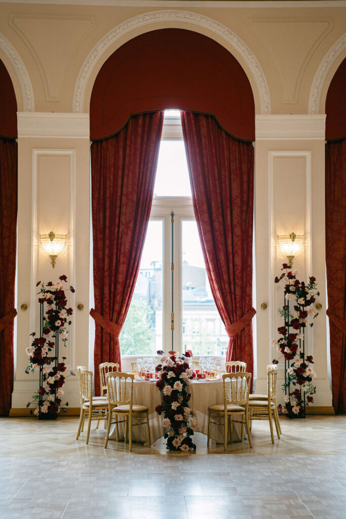 Intimate sweetheart table setup inside Cercle Cité Luxembourg, framed by a grand arched window with deep red draping and floral arrangements
