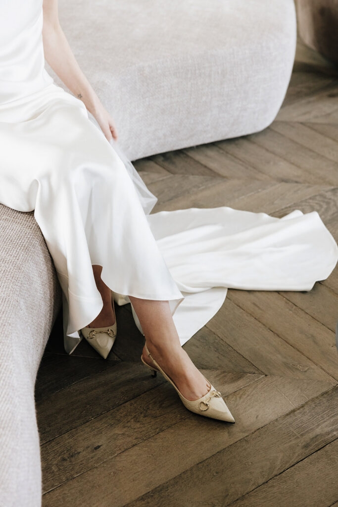 Bridal detail shot of gold heels and silk wedding gown on herringbone wood floor at Château de Mirwart Belgium