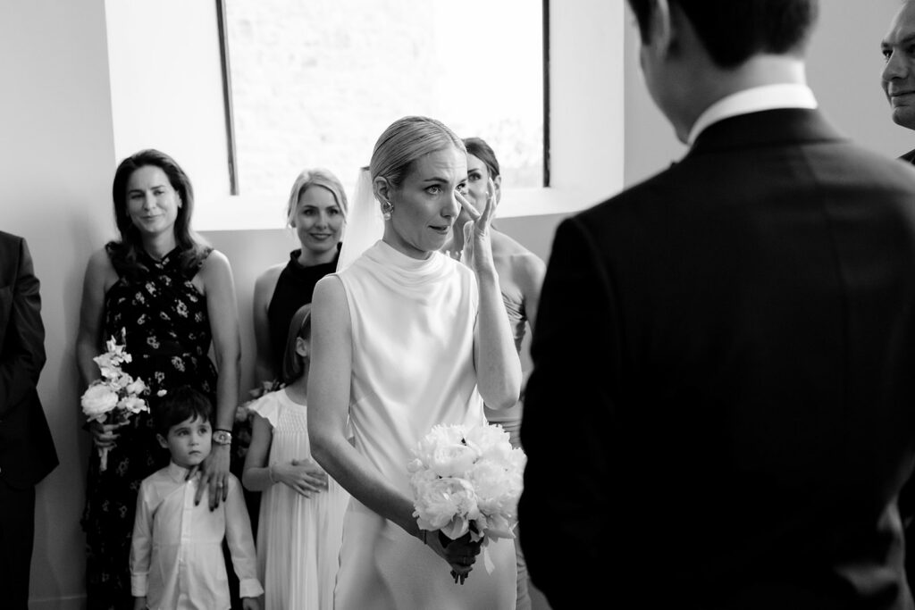 Bride Carla wiping away tears during her wedding ceremony at Château de Mirwart Belgium as groom Robin watches black and white