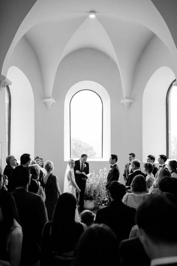Wide view of intimate wedding ceremony inside the gothic chapel at Château de Mirwart Belgium with guests gathered and white floral arrangements