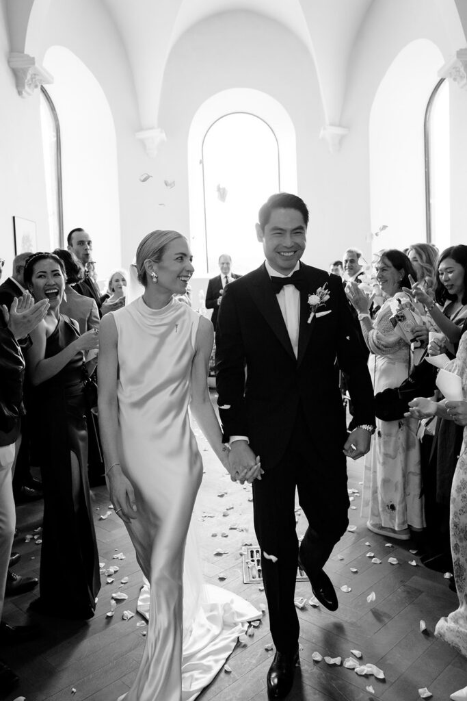 Carla and Robin walking back down the aisle as newlyweds at Château de Mirwart Belgium surrounded by cheering guests and rose petals black and white