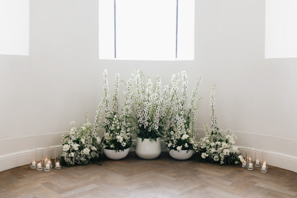 Wedding ceremony floral installation by Catherine Alves at Château de Mirwart Belgium featuring tall white delphiniums ranunculus and candlelight in white ceramic bowls on a herringbone wood floor