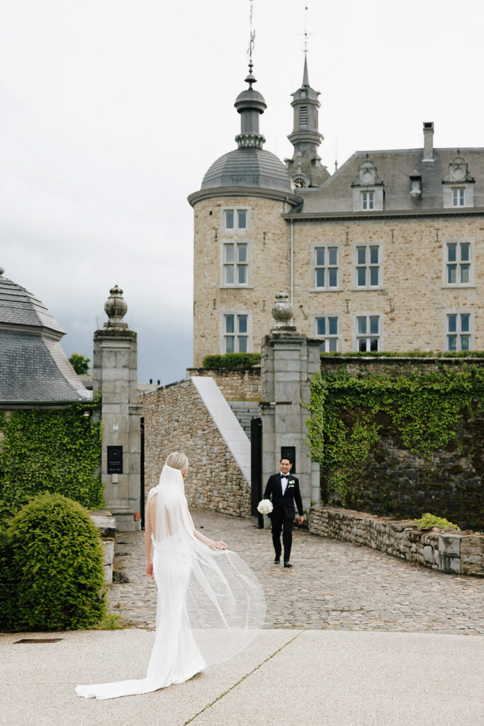 Bride and groom walking separately toward each other on the cobblestone bridge at Château de Mirwart Belgium with cathedral veil trailing