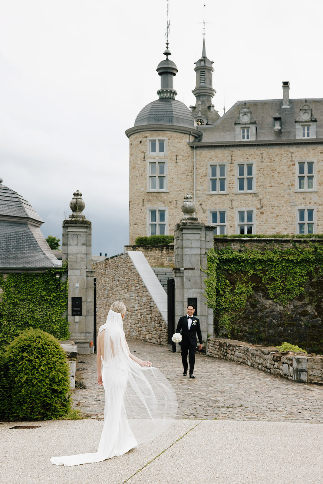 wedding portrait at the stone gate entrance of Château de Mirwart Belgium bride in silk gown with cathedral veil and groom in black tuxedo