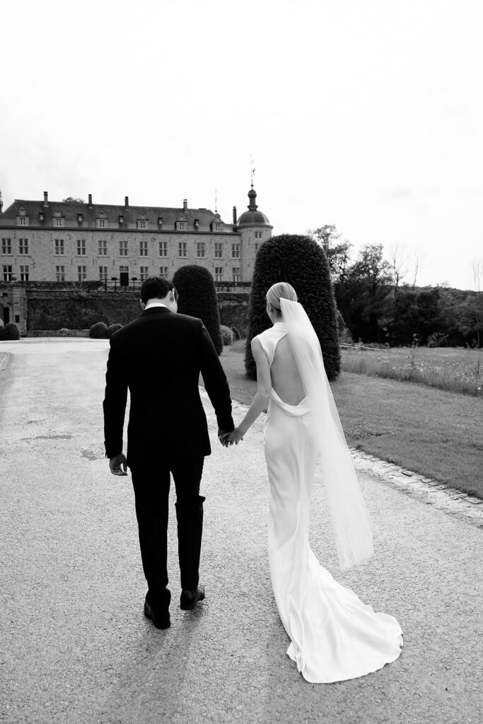 Black and white photo of Carla and Robin walking hand in hand toward Château de Mirwart Belgium showing the open back of Carla's silk bridal gown and veil