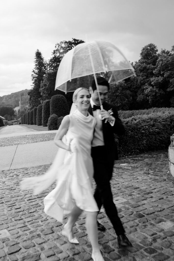 Carla and Robin laughing under a clear umbrella in the rain on the cobblestone grounds of Château de Mirwart Belgium black and white
