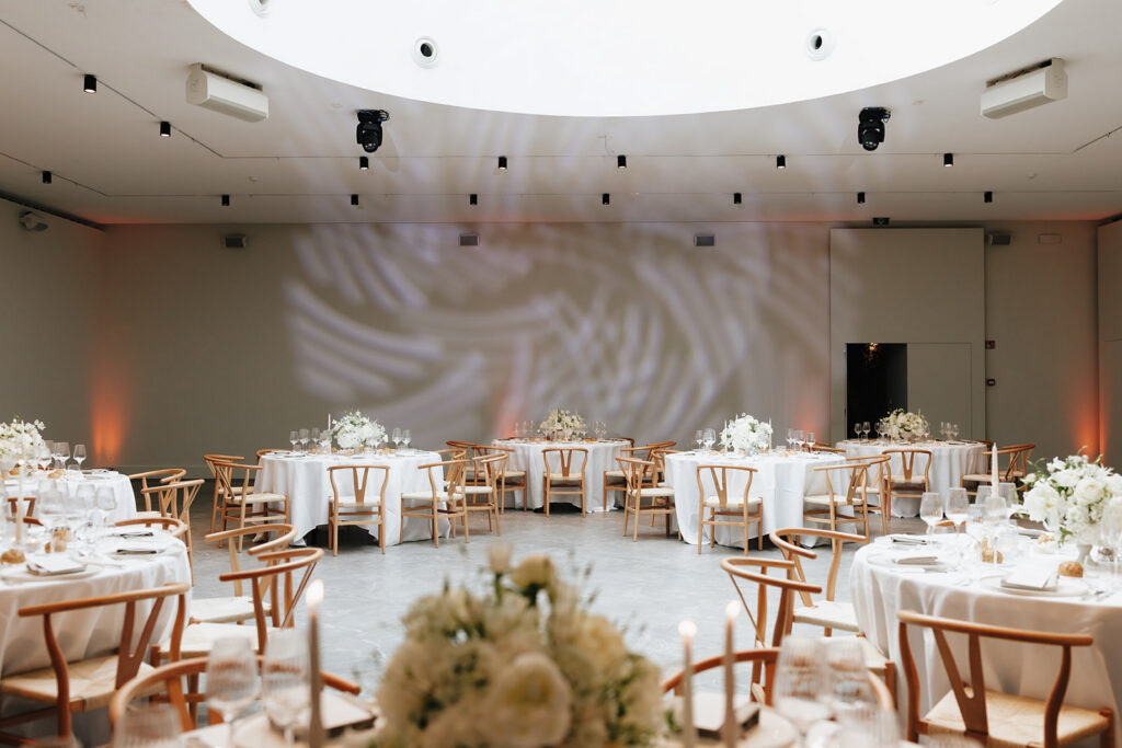 Wedding reception room at Château de Mirwart Belgium fully set with round tables wishbone chairs white floral centerpieces and dramatic projected leaf lighting on the walls