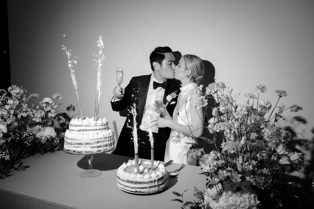 Carla and Robin kissing during their wedding cake cutting at Château de Mirwart Belgium with sparklers champagne and naked cakes surrounded by floral arrangements by Catherine Alves black and white