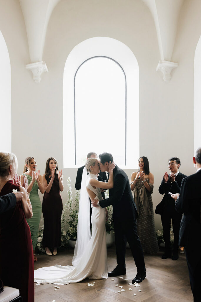 Carla and Robin sharing their first kiss as a married couple inside the chapel at Château de Mirwart Belgium surrounded by cheering guests with light streaming through a tall arched window