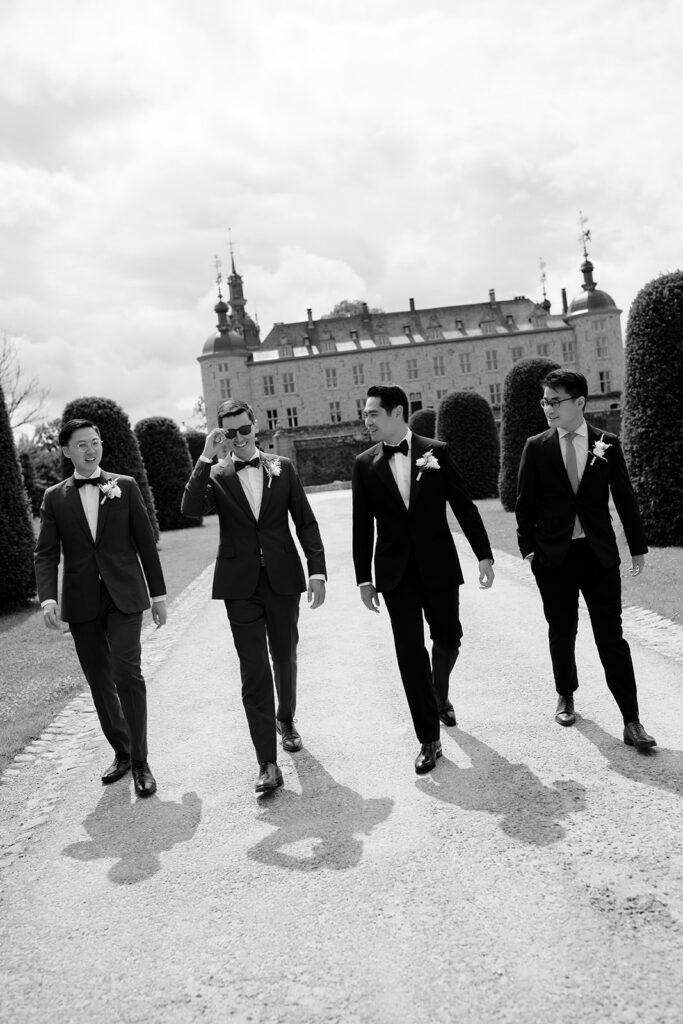 Black and white photo of groom Robin and groomsmen walking in black tuxedos on the grounds of Château de Mirwart Belgium