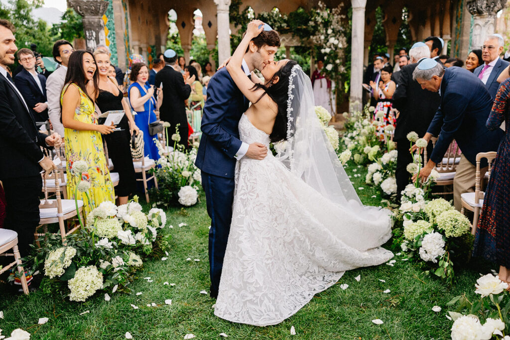 first kiss at their Jewish wedding ceremony in the gardens of Villa Cimbrone Ravello Italy surrounded by white hydrangea floral arrangements and cheering guests
