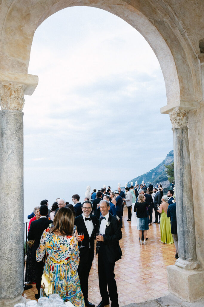Wedding cocktail hour on the terrace at Villa Cimbrone Ravello framed by a historic stone arch with guests mingling overlooking the Amalfi Coast and Mediterranean Sea