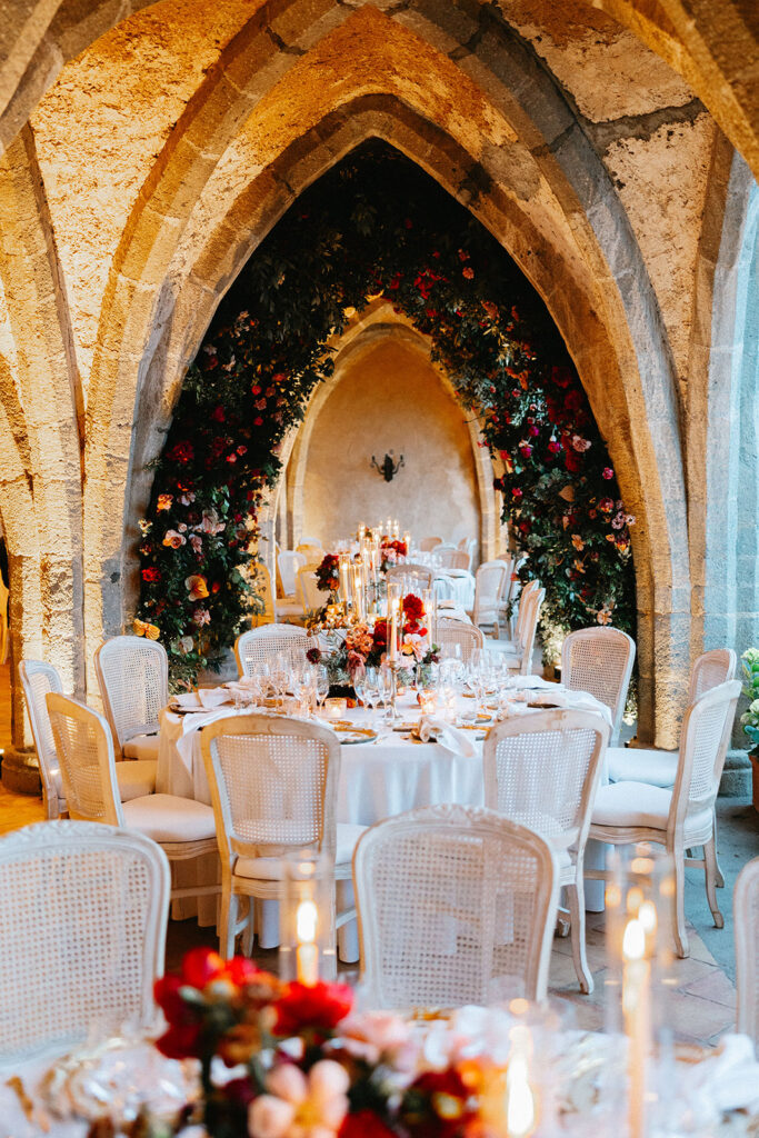 Wedding reception dinner table inside the Villa Cimbrone crypt Ravello with white cane chairs red and blush floral centerpieces taper candles and a lush floral arch framing the gothic stone alcove