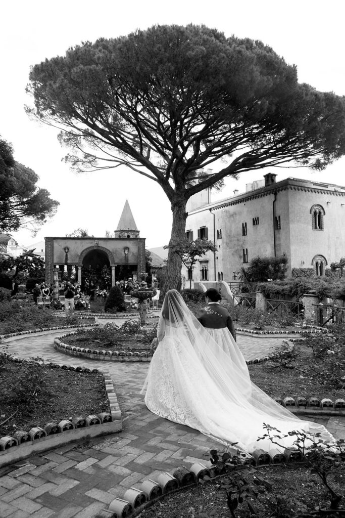 Black and white photo of bride walking toward her Jewish wedding ceremony in the gardens of Villa Cimbrone Ravello Italy with cathedral veil trailing behind her