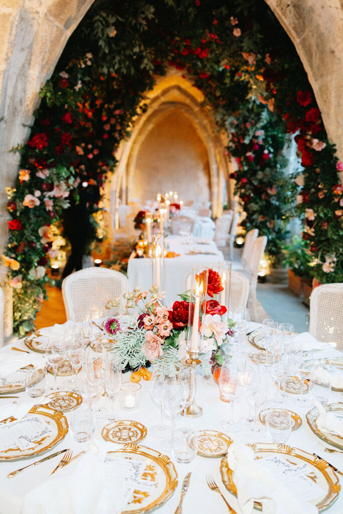 Luxury wedding reception dinner table inside the Villa Cimbrone crypt Ravello with gold rimmed china floral centerpieces and a dramatic floral arch installation in the background