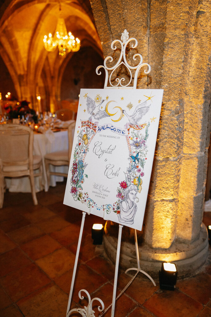Hand painted wedding welcome sign by the bride displayed on a white iron easel inside the candlelit crypt at Villa Cimbrone Ravello Italy