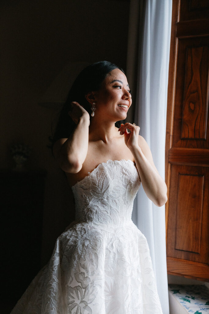 Bride Crystal putting on her earrings in her getting ready suite at Villa Cimbrone Ravello Italy smiling in her lace wedding gown
