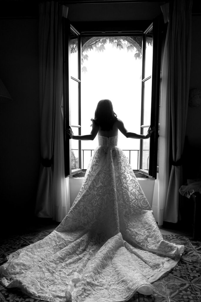 Black and white silhouette of bride in lace wedding gown with cathedral train standing at an open window at Villa Cimbrone Ravello Italy
