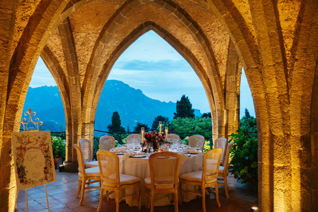Villa Cimbrone crypt wedding reception table framed by gothic stone arches with a sweeping view of the Amalfi Coast mountains at dusk through the open arch