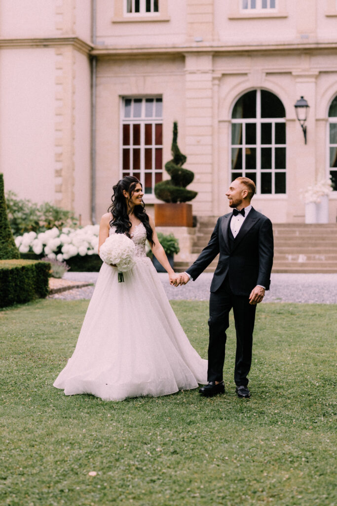 Bride and groom holding hands on the lawn in front of Château du Bois d'Arlon Belgium with white floral arrangements framing the château entrance