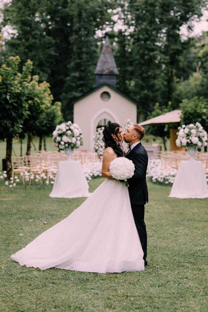 Bride and groom sharing a romantic moment on the lawn at Château du Bois d'Arlon Belgium with the chapel and white floral aisle in the background