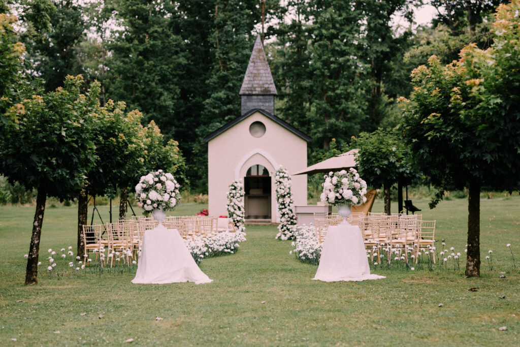 Wide view of outdoor wedding ceremony setup at Château du Bois d'Arlon Belgium with the chapel white carnation lined lawn gold Chiavari chairs and large floral urn arrangements surrounded by forest