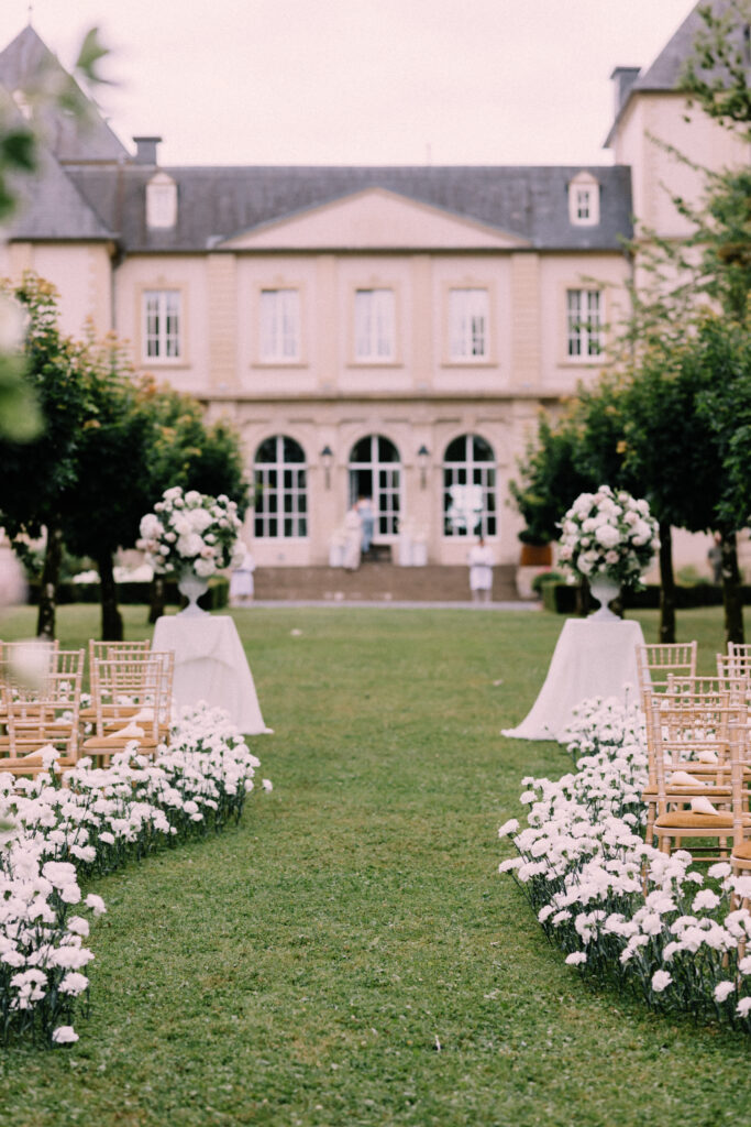 Outdoor wedding ceremony setup at Château du Bois d'Arlon Arlon Belgium with white floral aisle arrangements urns and gold Chiavari chairs on the grand lawn