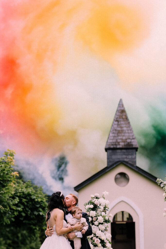 Bride and groom sharing their first kiss with their child during the wedding ceremony at Château du Bois d'Arlon Belgium with colorful fire smoke effect by Artifices.lu filling the sky above the chapel