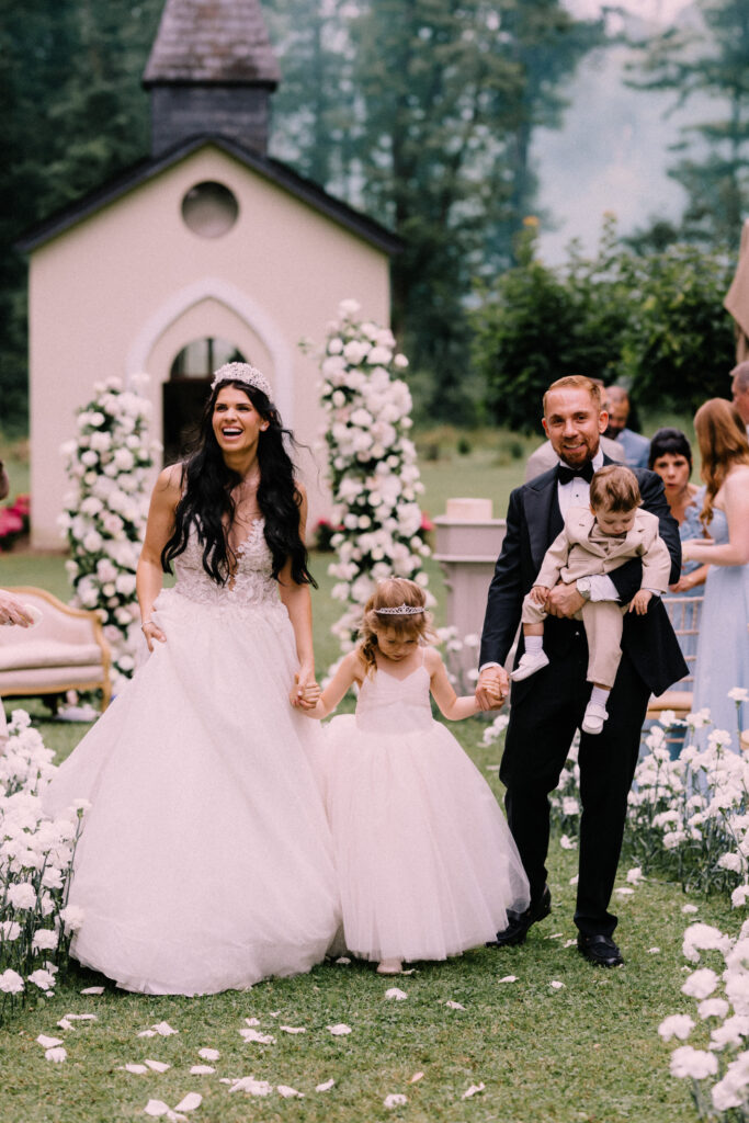 Bride groom and their two children walking back down the aisle after their wedding ceremony at Château du Bois d'Arlon Belgium with white floral arch and colorful smoke in the background