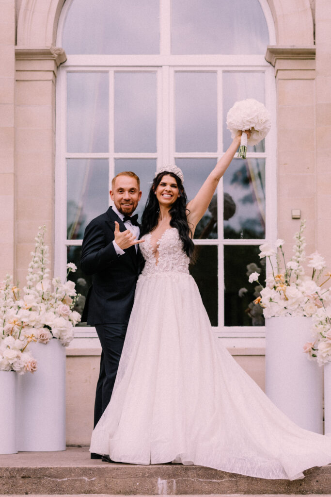 Bride raising her bouquet in celebration with groom after their wedding ceremony at Château du Bois d'Arlon Belgium in front of white floral arrangements and the château windows