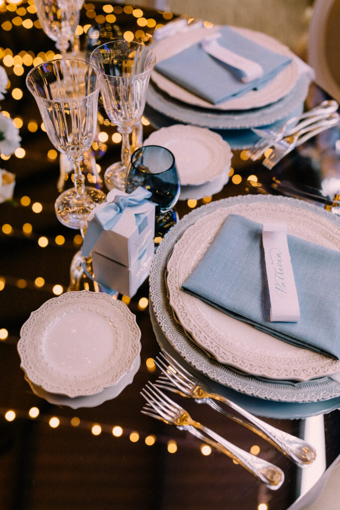 Wedding reception place setting detail at Château du Bois d'Arlon Belgium with blue linen napkin layered white china gold cutlery crystal glassware and fairy lights