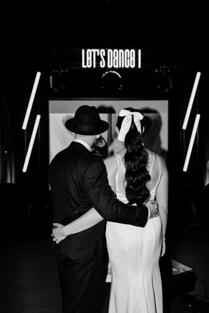 Bride in her second dress with white bow and groom in fedora hat walking hand in hand through a neon lit doorway reading Let's Dance at the Château du Bois d'Arlon wedding party in Belgium black and white