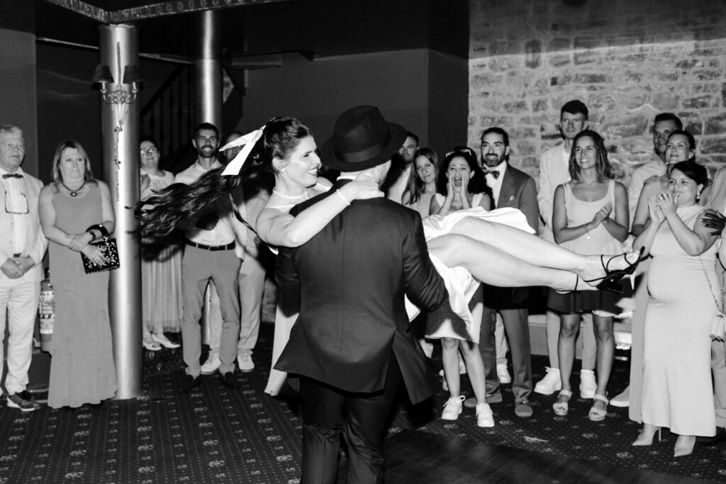 Black and white photo of groom lifting bride during the wedding party at Château du Bois d'Arlon Belgium with guests cheering and clapping around them