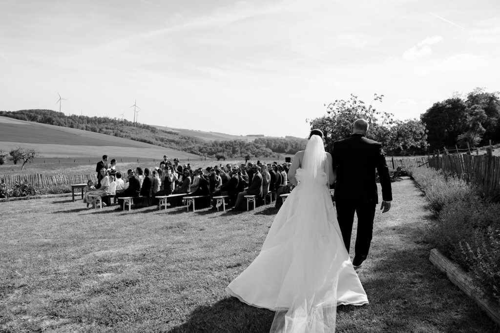 Hazelnut House 1851 Luxembourg wedding processional in black and white, the bride and her father walking toward the seated guests with her long train trailing across the lawn, rolling countryside and hilltop wind turbines beyond