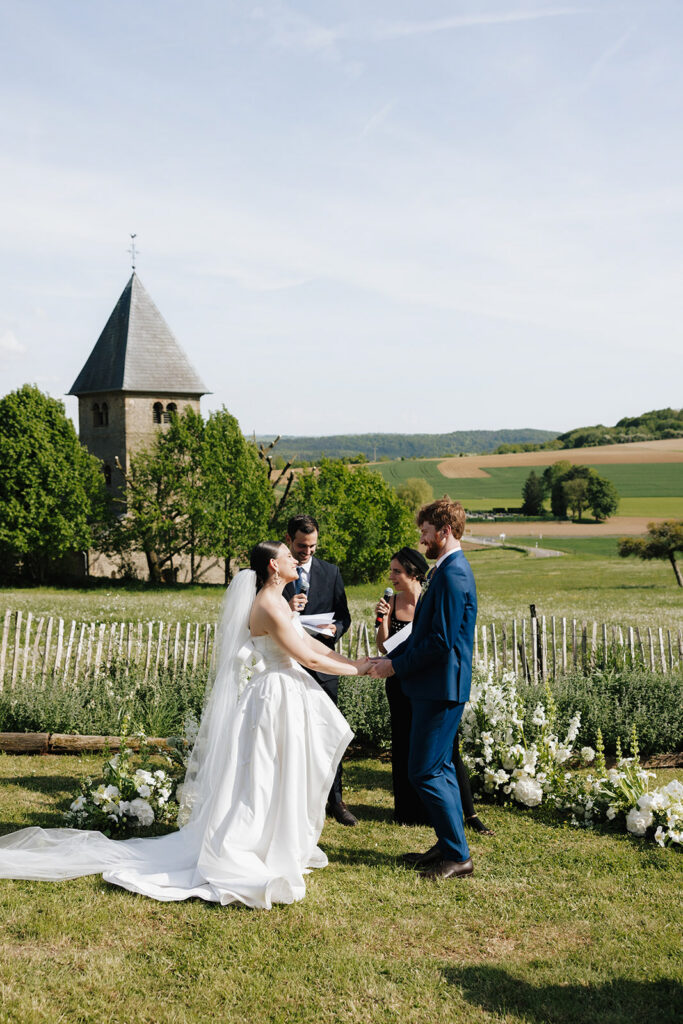 Hazelnut House 1851 Luxembourg wedding ceremony held outdoors, bride in a strapless gown and veil holding hands with her groom in a navy suit before a historic stone church tower and rolling countryside, white floral arrangements lining the ceremony space