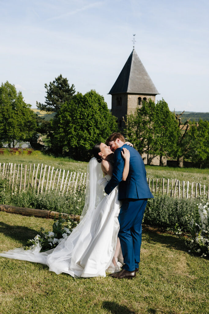 Hazelnut House 1851 Luxembourg wedding portrait, the bride and groom sharing a kiss on the lawn before the historic stone church tower and rustic fence lined with florals