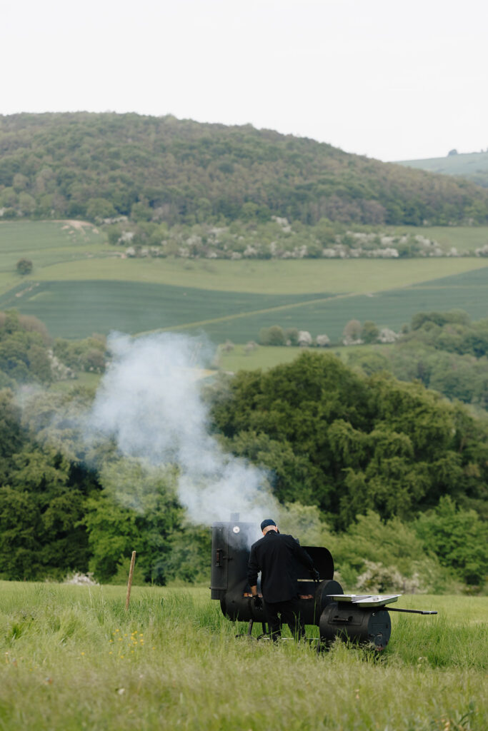 Hazelnut House 1851 Luxembourg wedding open air catering, a chef tending a smoking grill in the meadow with rolling green hills and vineyards beyond