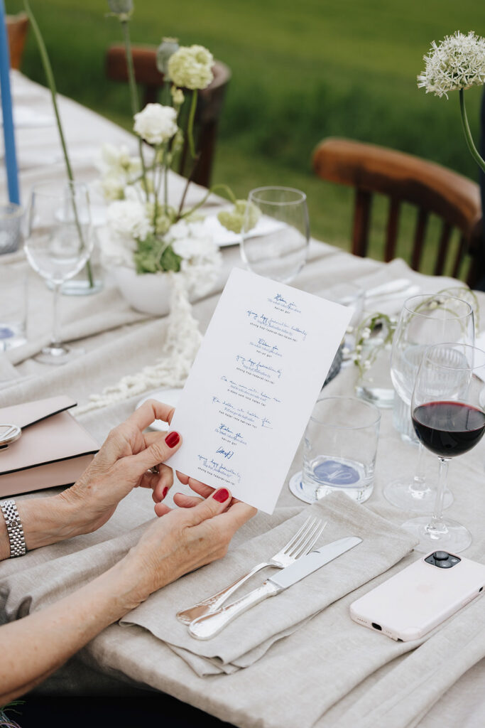 Hazelnut House 1851 Luxembourg wedding reception detail, a guest holding the printed menu at a table set with white florals, red wine, and refined place settings